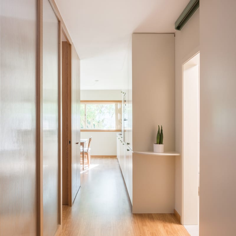 Corridor with reeded-glass sliding panels, oak floor and a curved shelf, leading to a bright window.